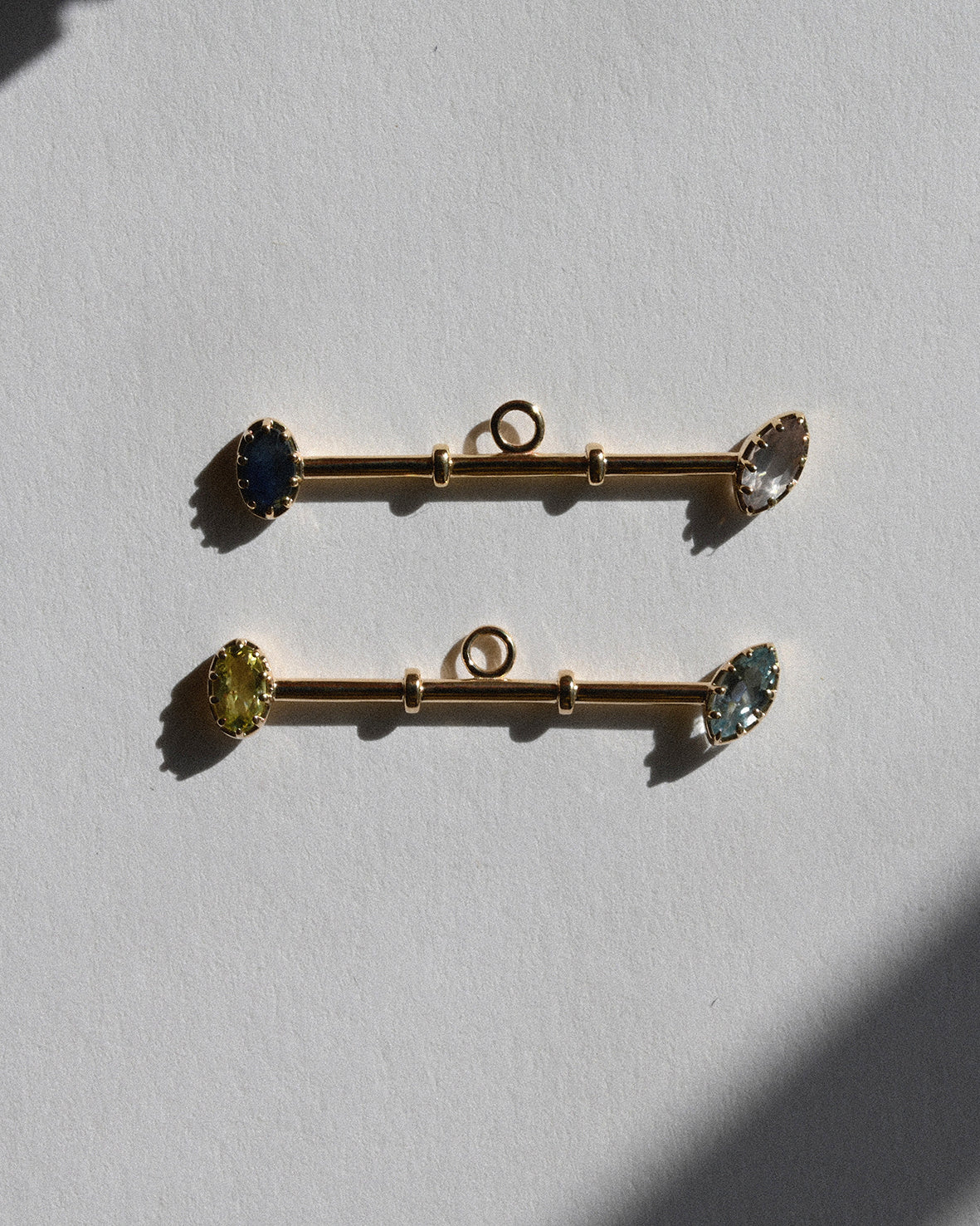 Two gold bar pendants with gemstones on a light gray background