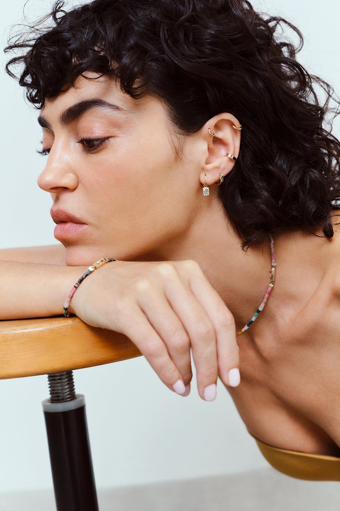 A close-up lifestyle shot of a model with dark curly hair resting her head on a wooden table, showcasing a delicate beaded bracelet on her wrist and a matching beaded necklace around her neck.