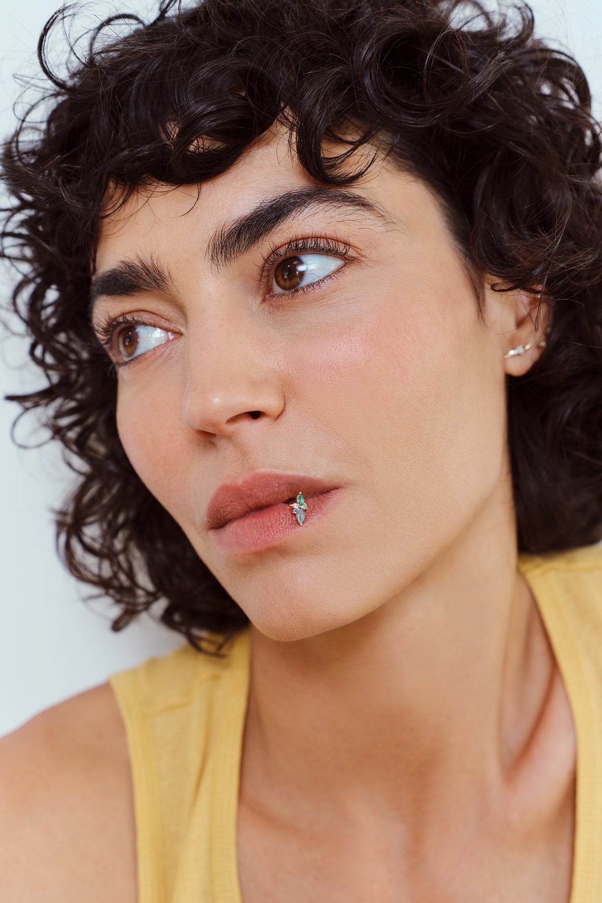 A close-up portrait of a woman with short curly dark hair wearing a yellow top, looking to the side. She is wearing a small metier gold hoop earring with a kaleido gemstone  stud on her lower lip.