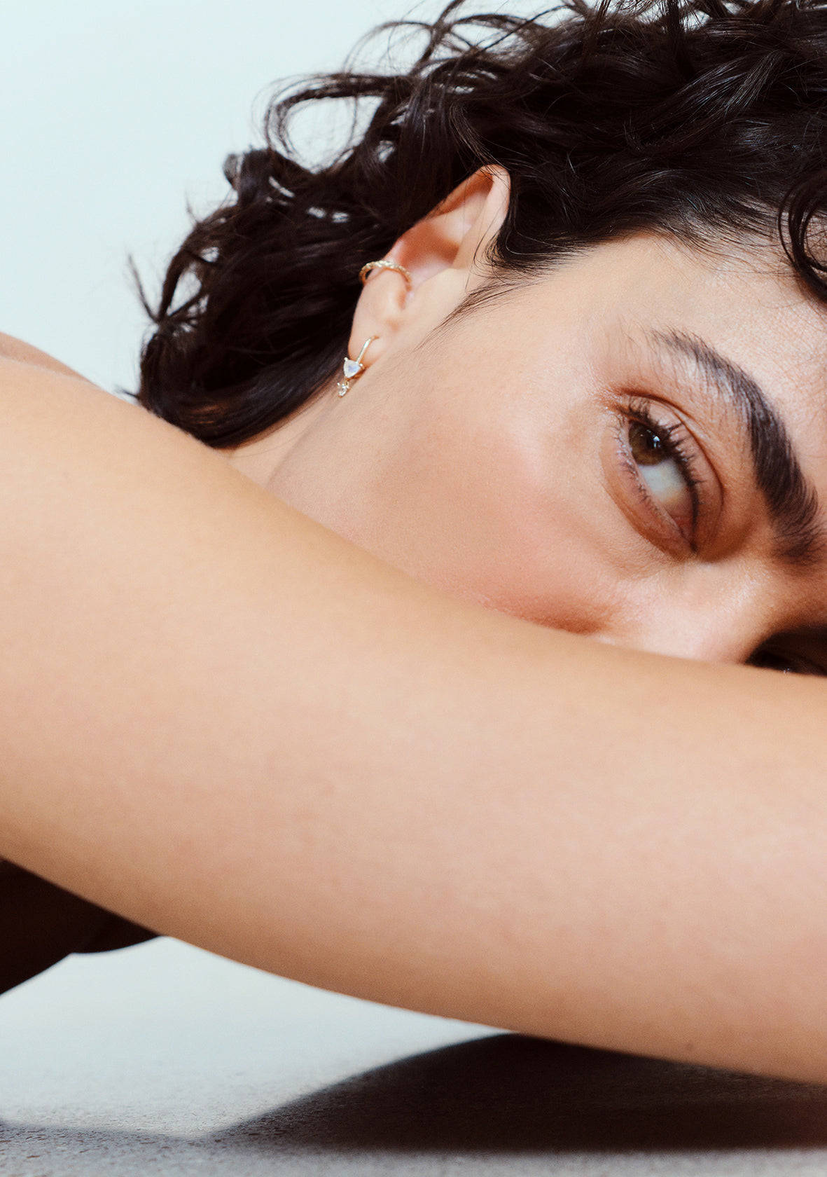 A close-up  shot of a person with dark curly hair resting their head on their arm, wearing a az trillion gold hoop earring in my métier by tomfoolery 