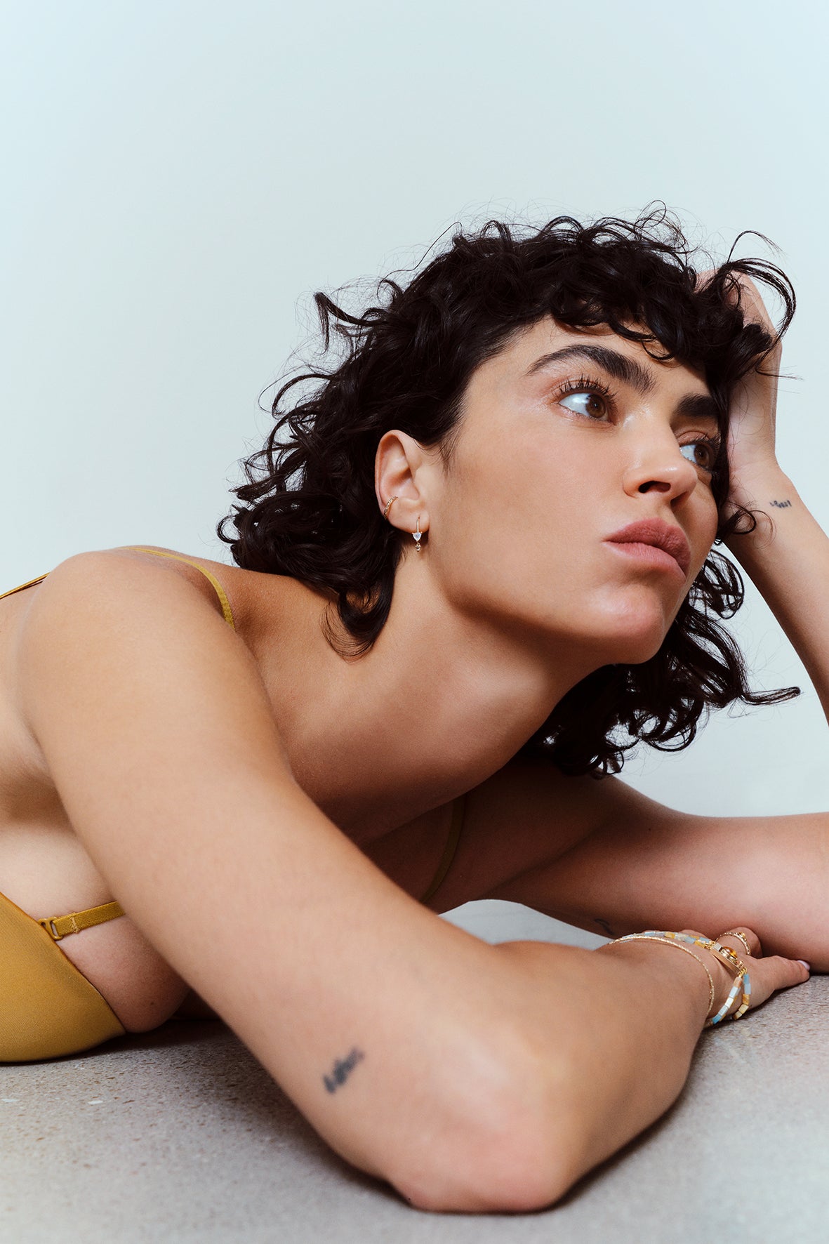 A woman with short curly dark hair lies on a light gray surface, wearing a mustard yellow bikini top. She is looking upward and to the side, with her hand resting near her head wearing metier by tomfololery az trillion drop